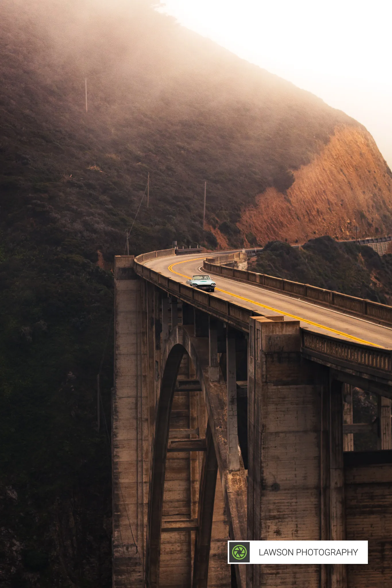 Light blue convertible driving across Bixby Creek Bridge in Big Sur under misty golden-hour light.