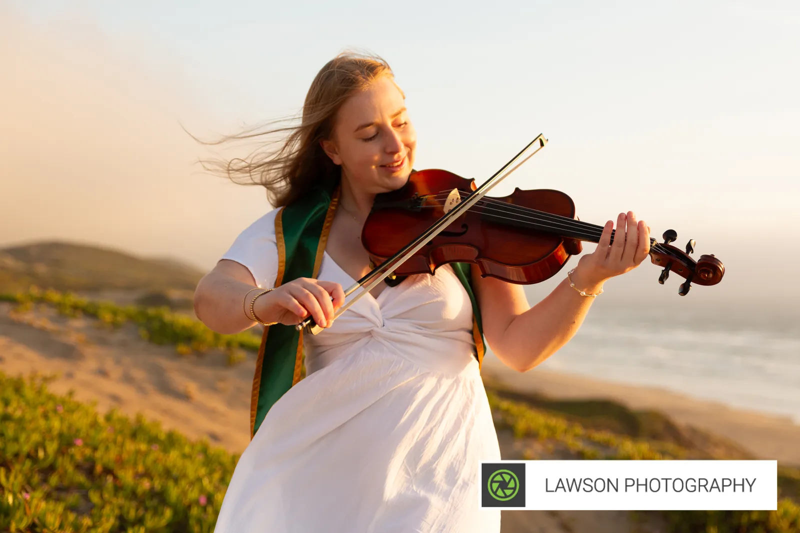 Young woman playing viola on a coastal dune at golden hour, eyes closed, hair blowing across her face, green sash over a white dress, ocean behind her.