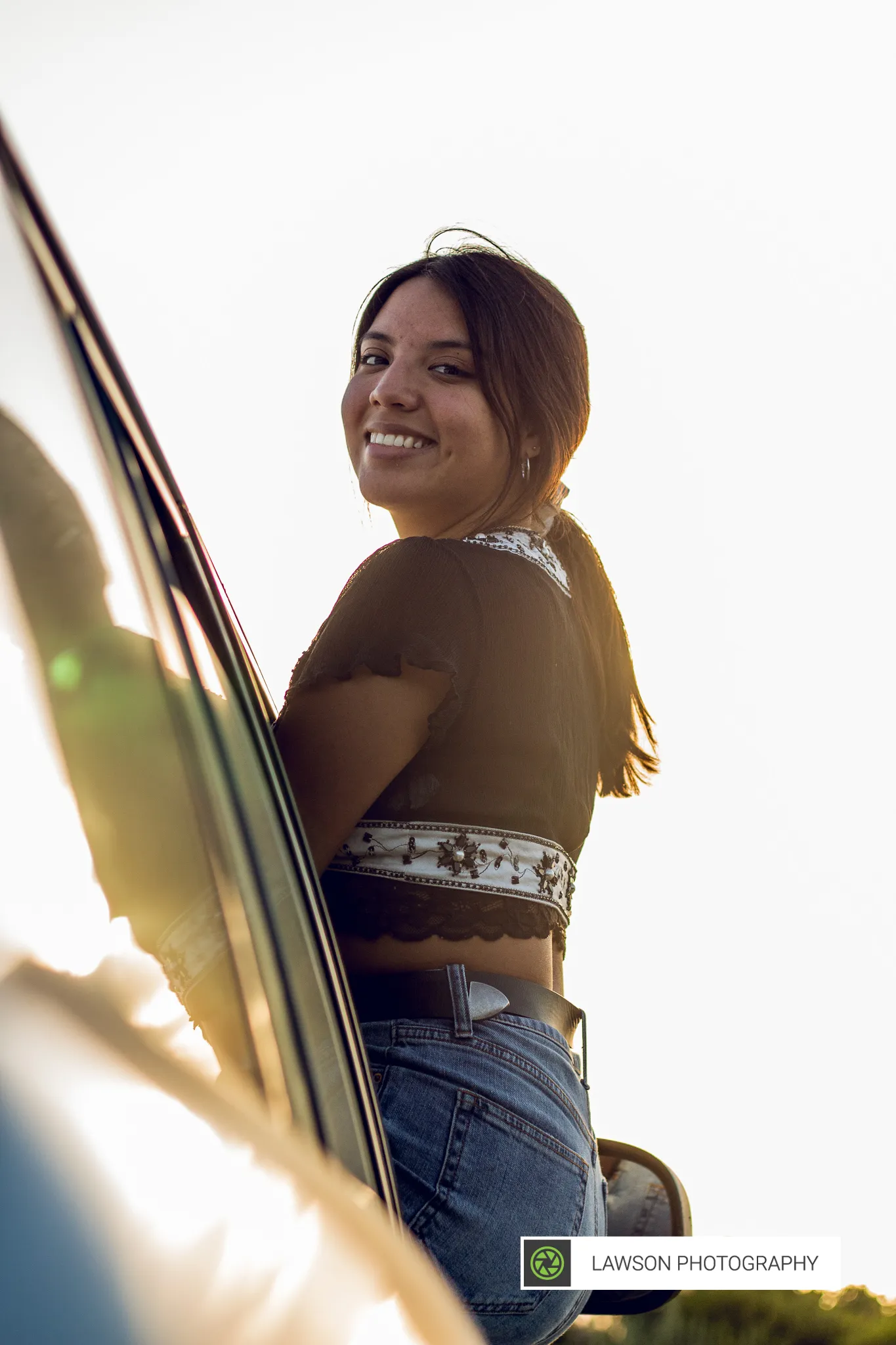 Young woman leaning from a car window, smiling over her shoulder in bright backlit sunlight.