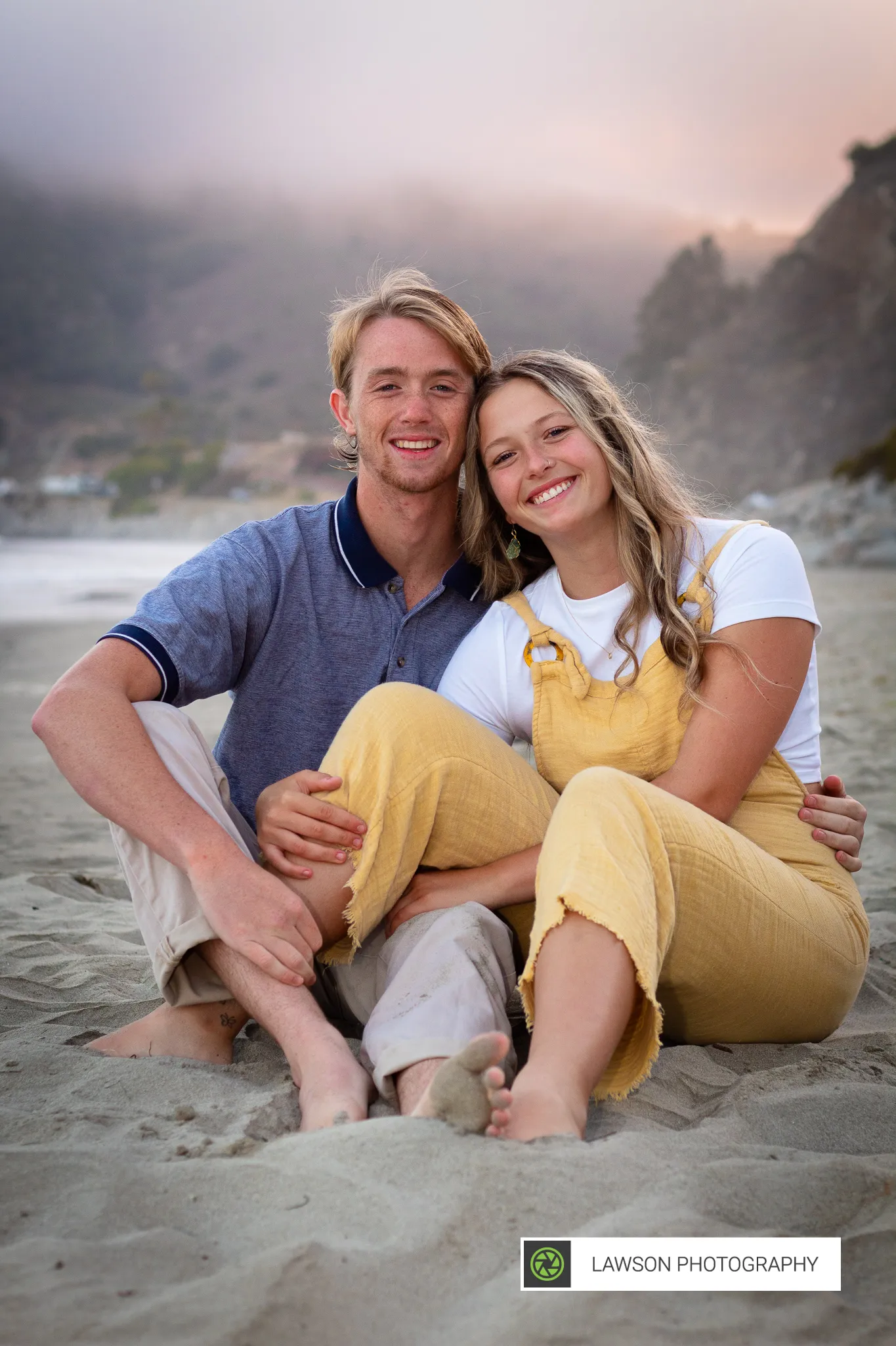 Young couple sitting barefoot on the sand, smiling together with misty coastal hills behind them.