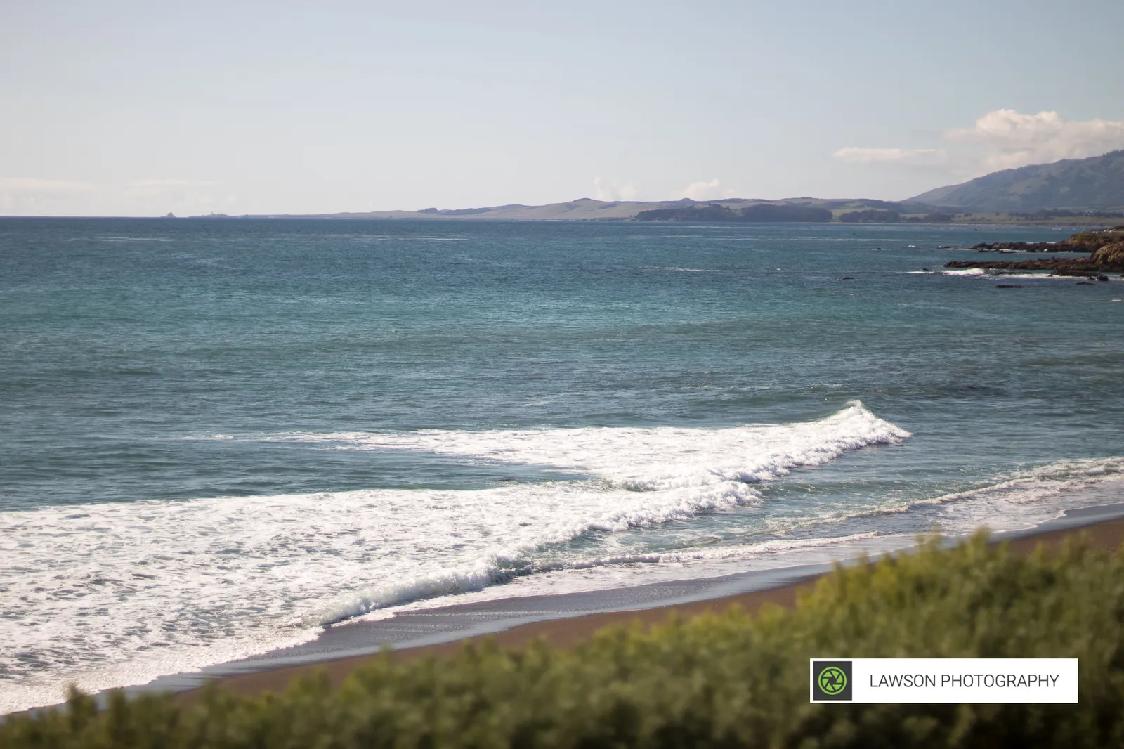 Elevated view of a dark-sand beach where a white V-shaped wave breaks toward distant coastal hills.