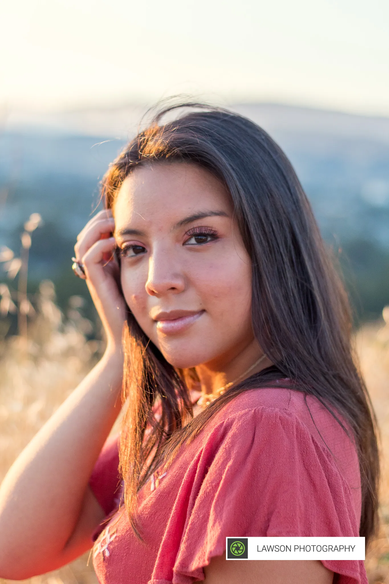 Close portrait of a young woman brushing dark hair aside in coral blouse light, with golden hillside bokeh behind her.