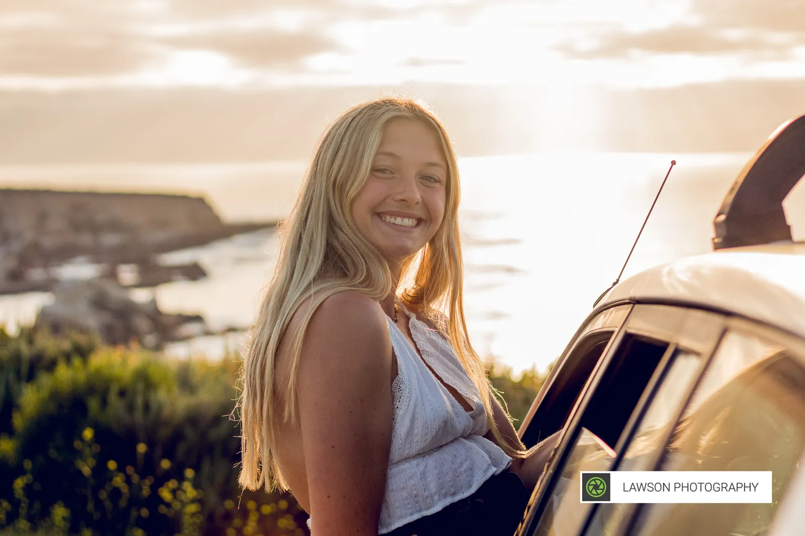 Smiling young woman in a white sleeveless top leaning by a white car with ocean cliffs glowing at sunset behind her.