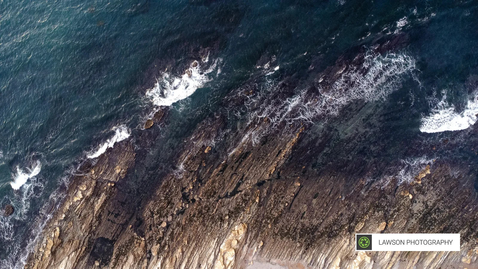 Top-down aerial of deep blue-green waves crashing on layered dark rocky shore.
